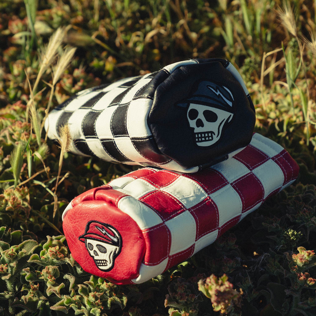 Two checkered skate pads, one black and white with a skull design, and the other red and white, are displayed on a grassy surface.